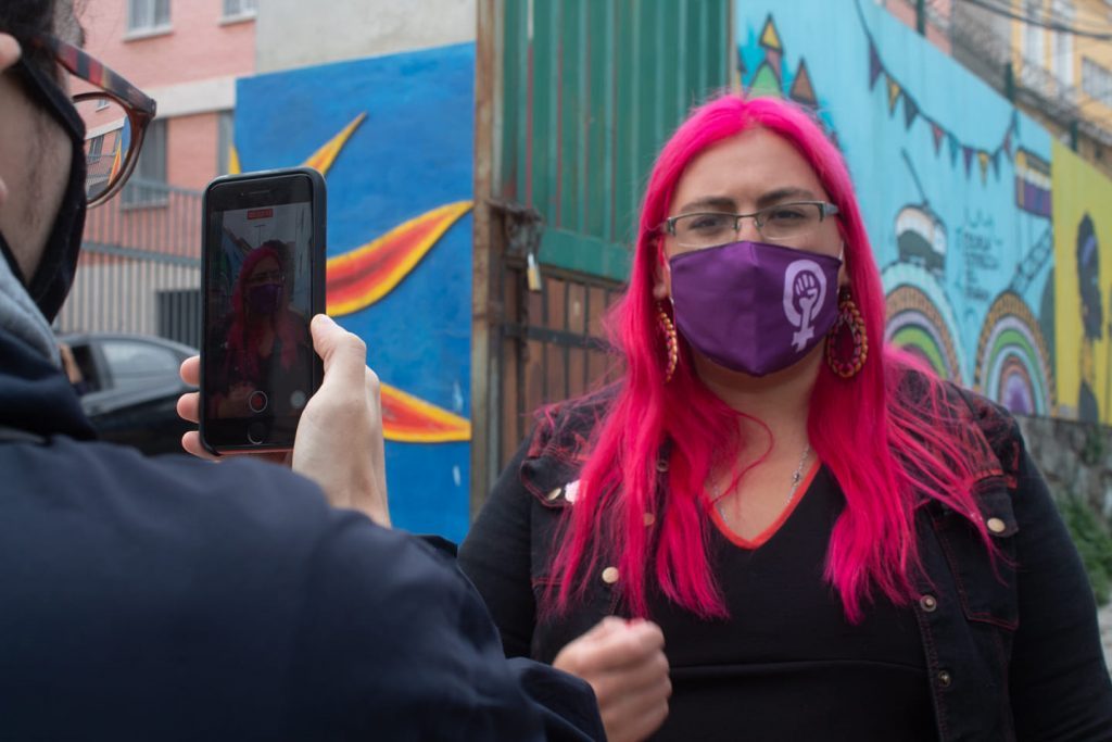Constanza Valdés con cabello rosa y mascarilla púrpura siendo fotografiada frente a mural callejero colorido