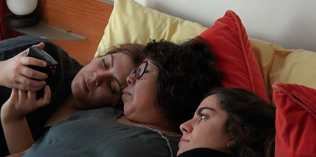 Tres personas acostadas en una cama viendo una película, con almohadas rojas y amarillas, en escena íntima