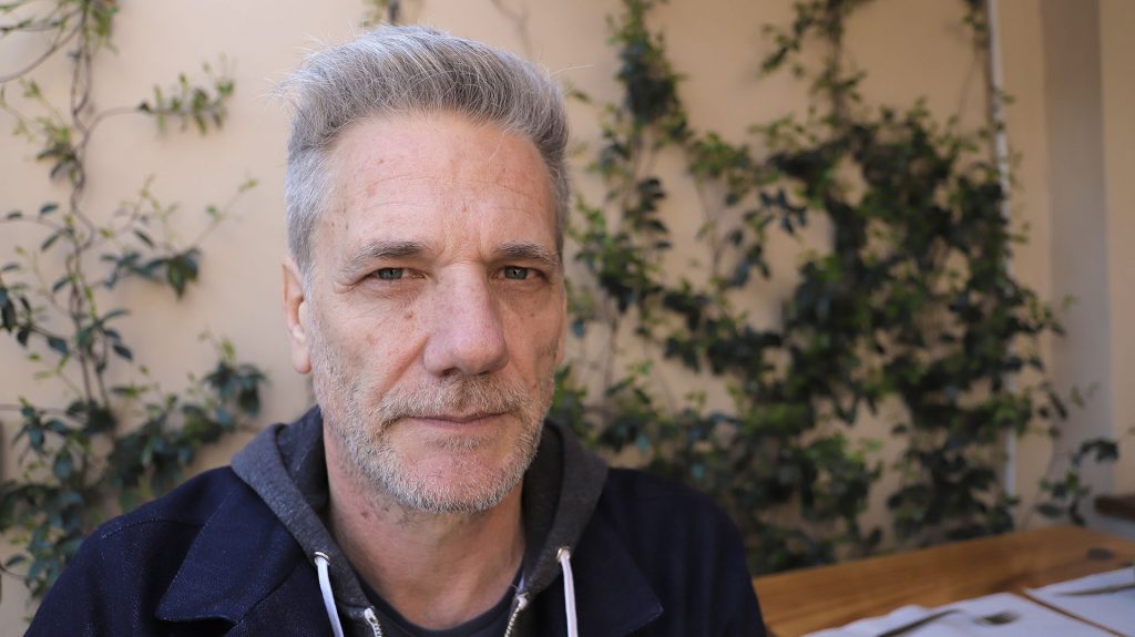 Alan Pauls, escritor argentino de cabello gris, viste chaqueta azul oscura sobre fondo con enredaderas