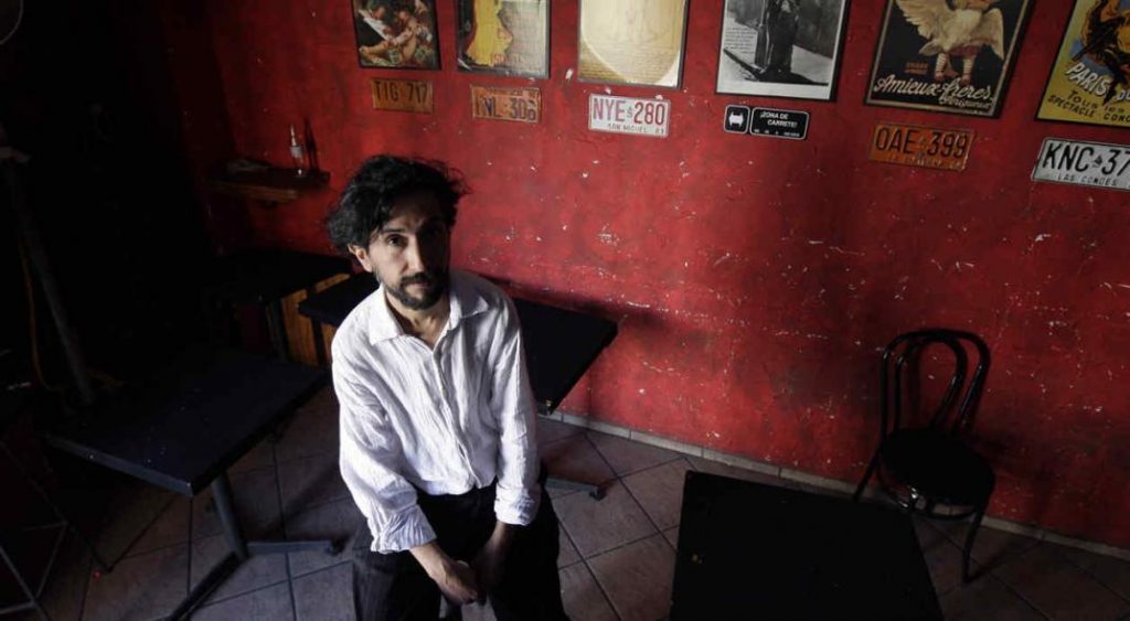 Leonardo Sanhueza sentado en una silla contra pared roja, en ambiente interior oscuro, mirando hacia la cámara