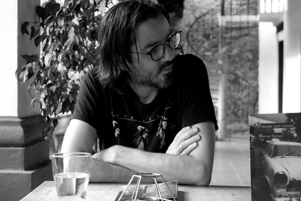 Manuel Illanes, hombre de cabello oscuro y gafas, sentado ante una mesa con vasos, brazos cruzados, en blanco y negro