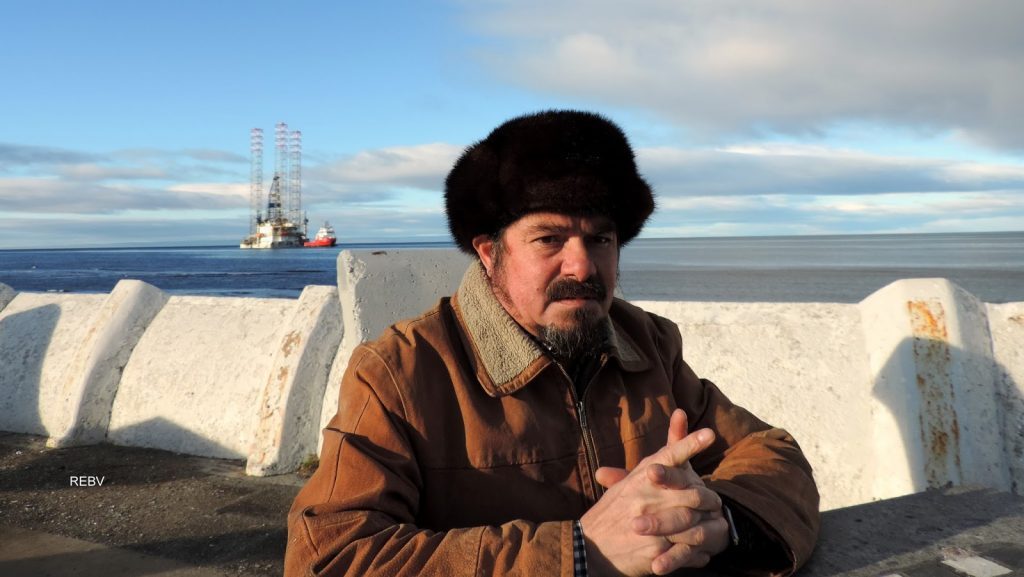Pavel Oyarzún en primer plano con gorro negro, fondo de playa y barco petrolero al horizonte