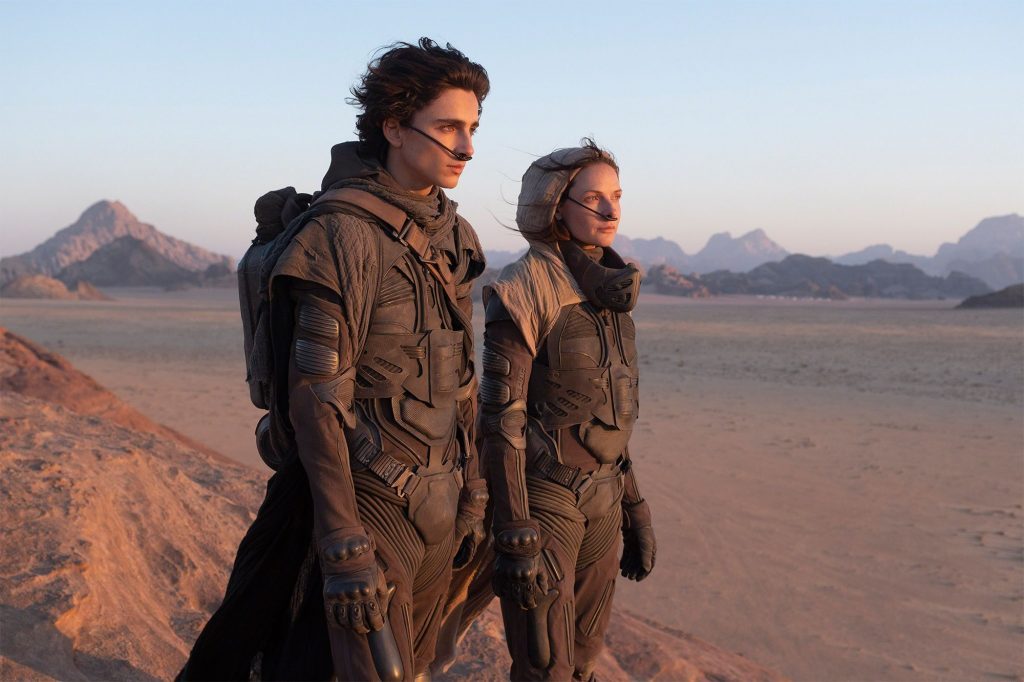 Dos personajes vistiendo trajes de supervivencia en un vasto desierto árido con montañas al fondo