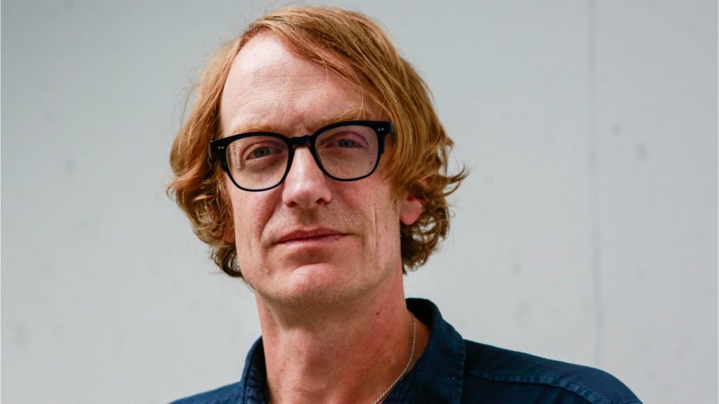 Patrick deWitt, escritor canadiense con gafas y cabello rojo ondulado, retrato de estudio sobre fondo gris claro