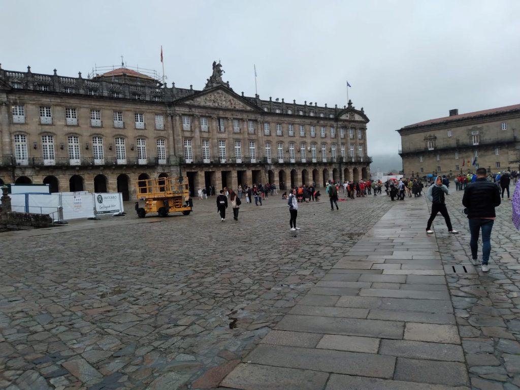 Plaza mayor europea con edificios históricos de piedra, personas caminando sobre adoquines grises en día nublado