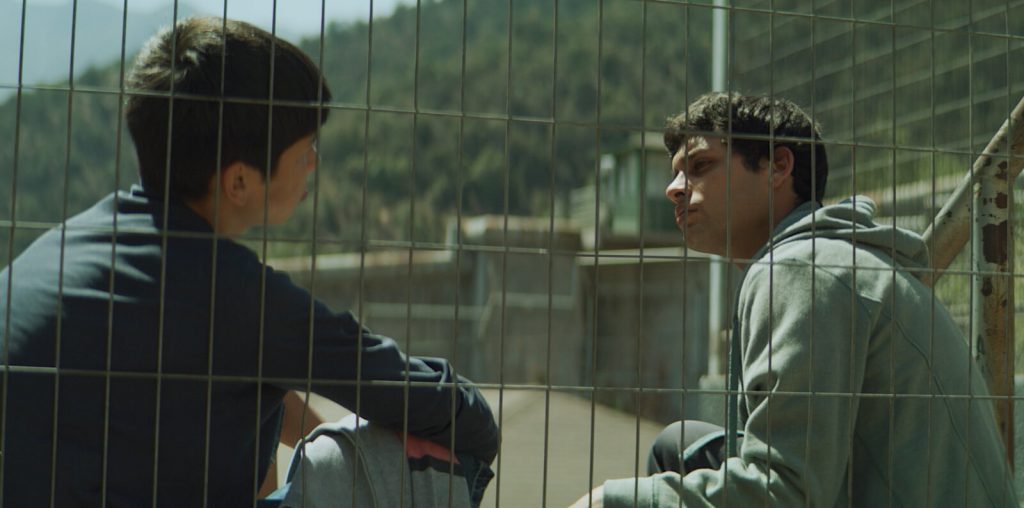 Dos jóvenes conversando frente a una cerca de metal en un entorno natural, escena de la película Mis hermanos sueñan