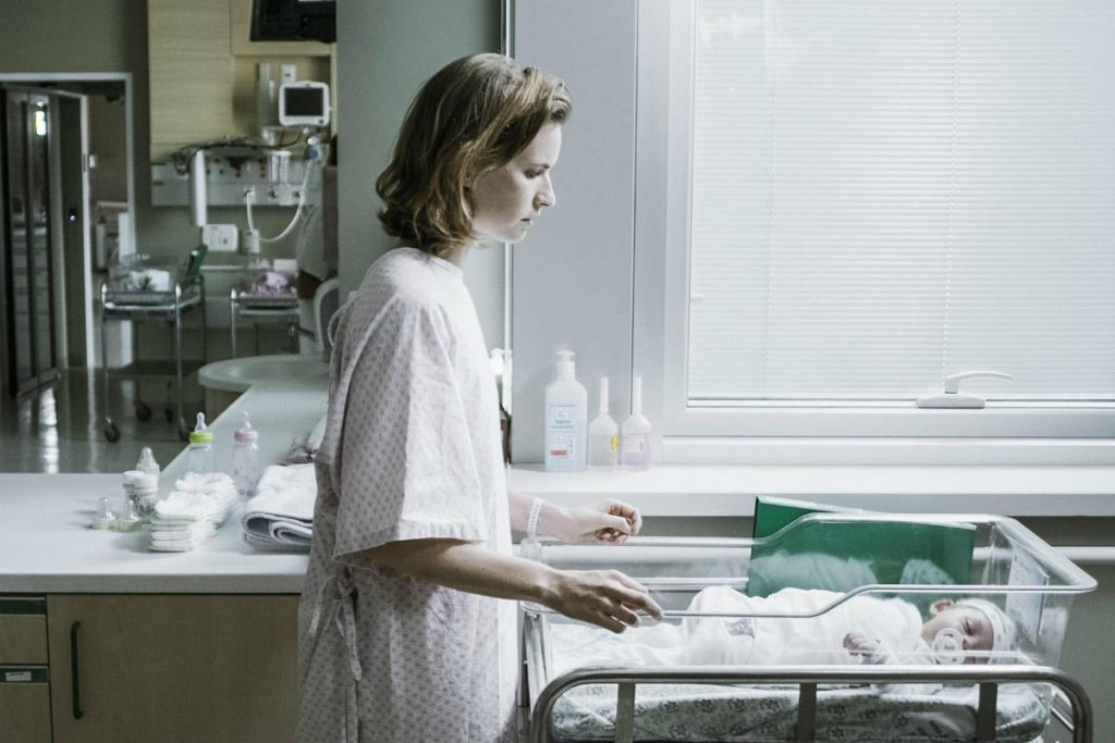 Mujer en hospital vistiendo bata de paciente, de perfil junto a una cama médica con equipos de monitoreo