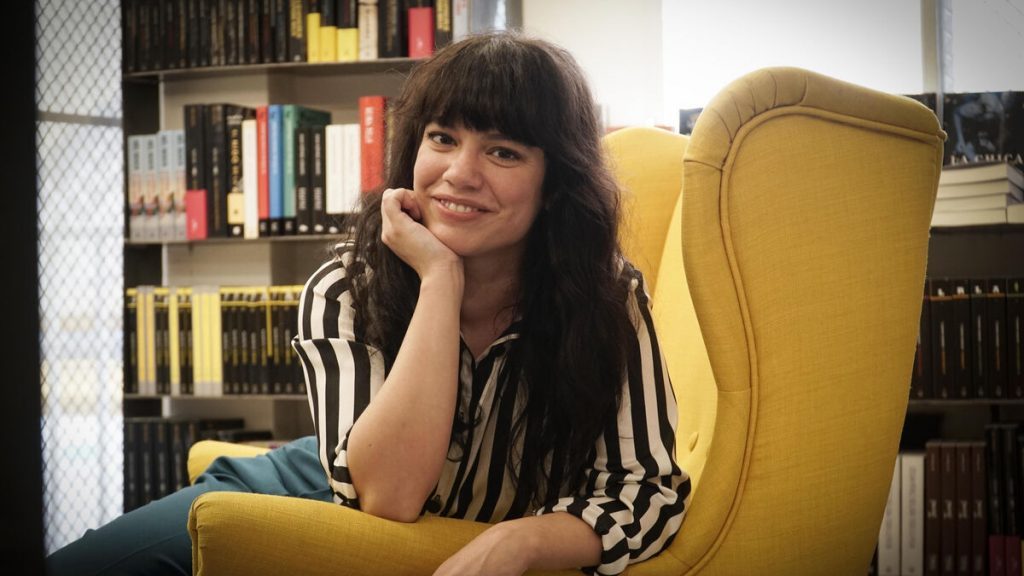 Elisa Victoria, escritora chilena, sentada en sillón amarillo sonriendo ante la cámara en su estudio con libros