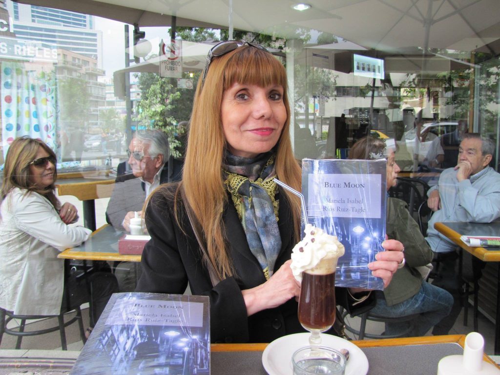 Mariela Ruiz Tagle sostiene libro azul en cafetería, sonriendo hacia cámara con ambiente de trabajo colaborativo