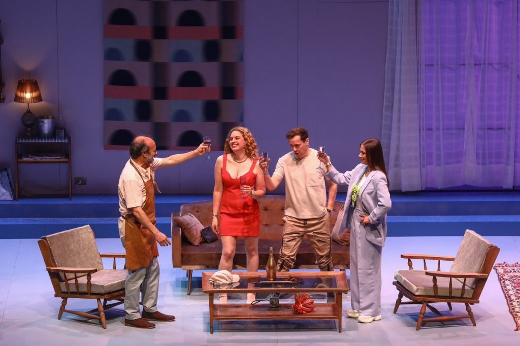 Cinco actores en escena de teatro moderno sobre plataforma elevada, con muebles de época y fondo iluminado en tonos