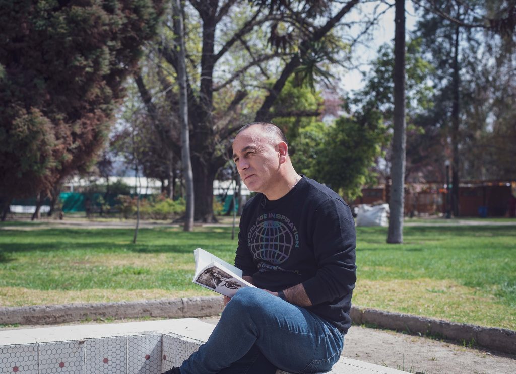 Hombre sentado en banco de parque sosteniendo un libro, con árboles de fondo y área verde