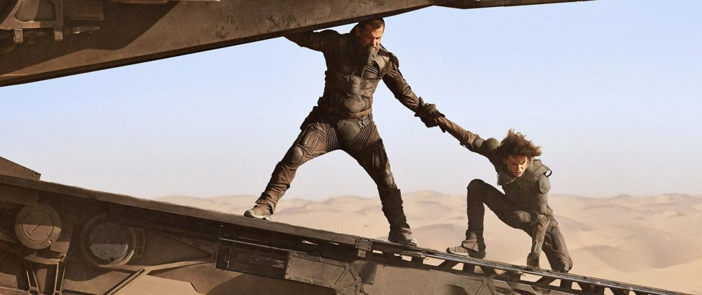 Dos personajes en túnica extendiendo la mano sobre una viga metálica en desierto árido con cielo claro