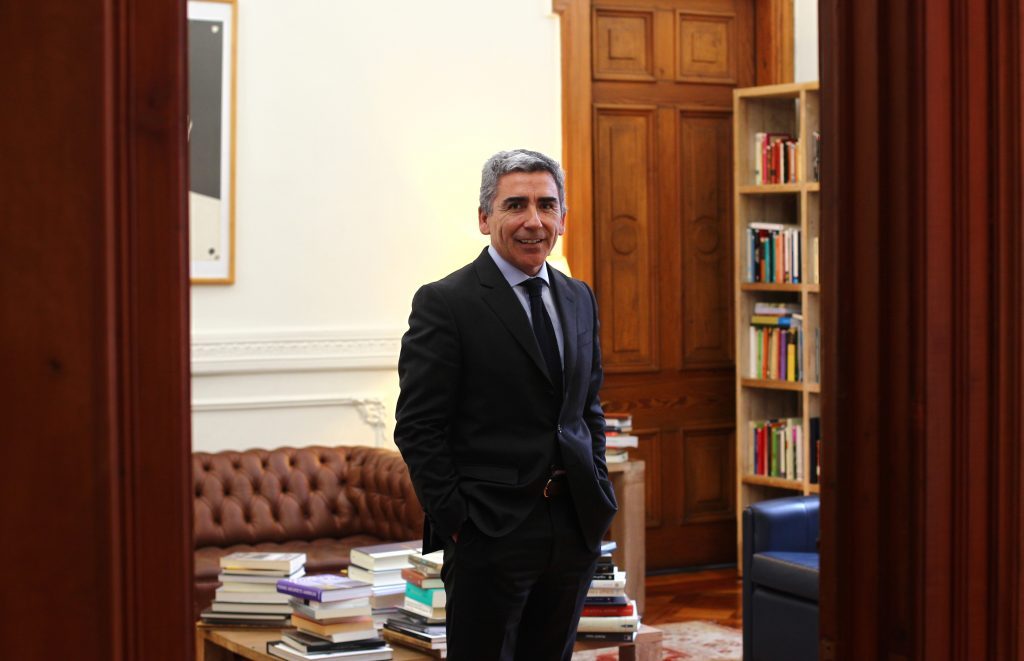 Hombre de mediana edad en traje oscuro y corbata azul dentro de una biblioteca, junto a estanterías con libros