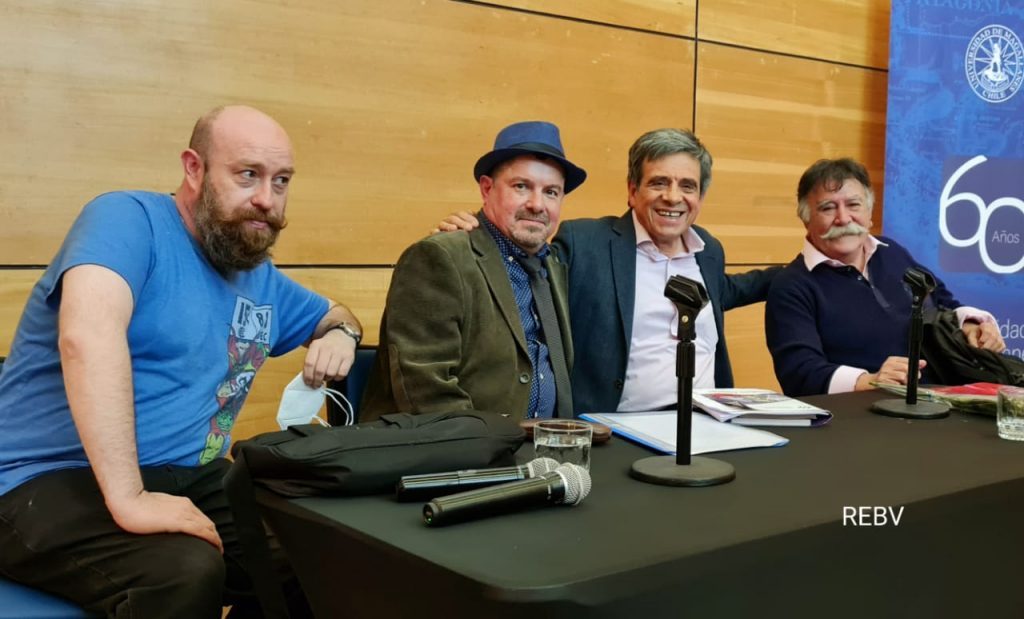 Cuatro hombres en panel de presentación de novela, uno con micrófono, sentados ante mesa con logotipo visible al fondo