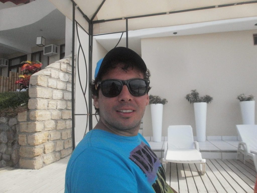 Hombre con gafas de sol y camisa azul en terraza con macetas blancas y vista a edificio de ladrillo
