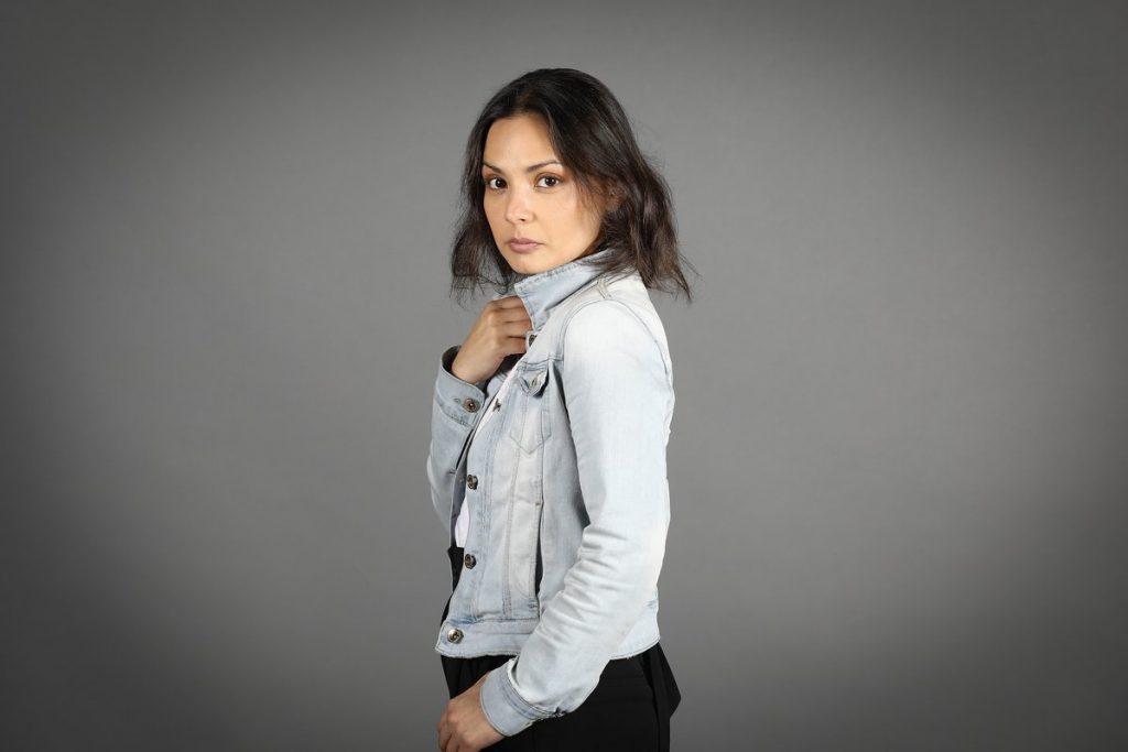 Mujer de cabello oscuro vuelteada hacia la cámara, vistiendo blusa blanca y chaleco gris sobre fondo neutro