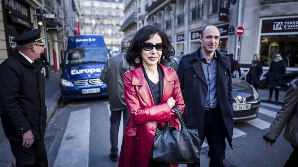 Yasmina Reza en una calle parisina, vistiendo abrigo rojo, rodeada de transeúntes y automóviles