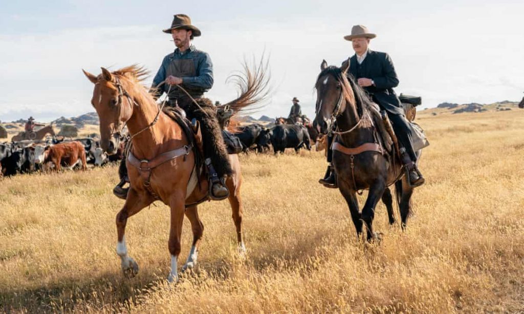 Dos vaqueros a caballo en la pradera árida, el de la izquierda monta un caballo marrón y viste camisa azul con sombrero