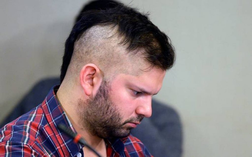 Hombre con barba y cabello oscuro, camisa a cuadros roja y azul, mirando hacia abajo en primer plano