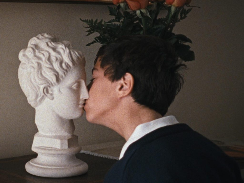 Hombre de perfil besando una estatua de yeso blanco clásica con plantas de fondo
