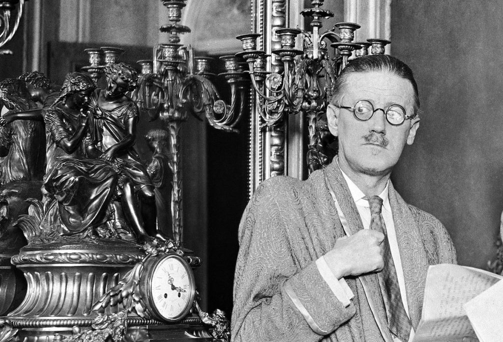 James Joyce con gafas sostiene papeles junto a un busto decorativo y maquinaria de escritura