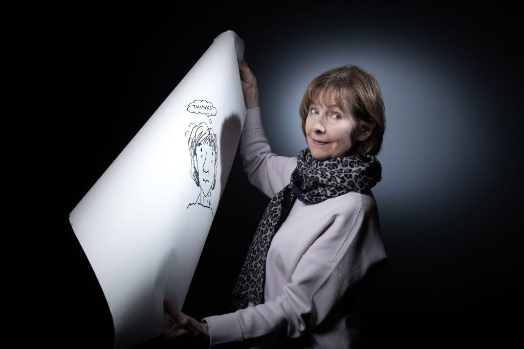 Posy Simmonds sostiene un cómic enrollado mientras posa contra un fondo gris oscuro en retrato profesional
