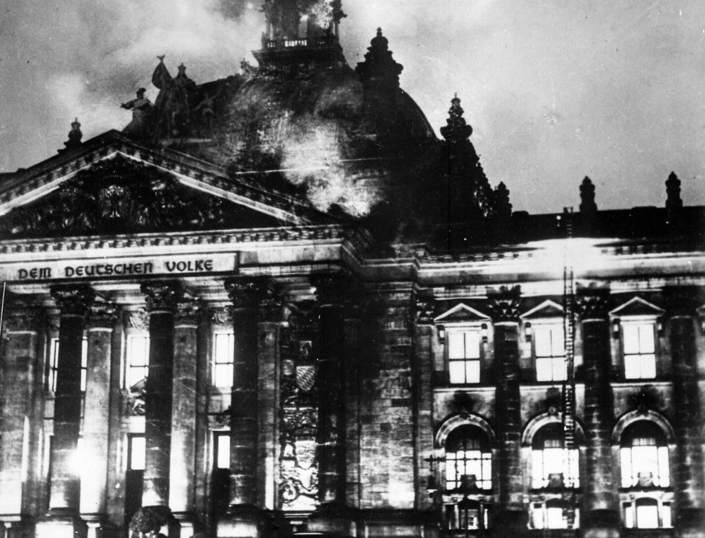 Edificio del Reichstag en llamas durante la noche, estructura neoclásica con columnas y cúpula ardiendo, humo denso
