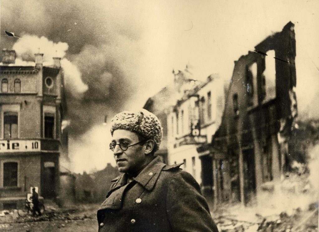 Vasili Grossman con gafas, de perfil, ante edificios en ruinas y humo de destrucción al fondo
