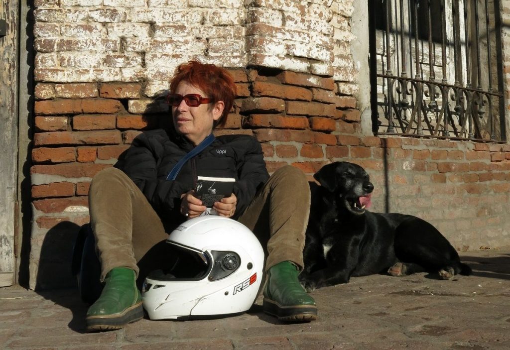 Cynthia Rimsky sentada contra pared de ladrillo con casco de moto blanco y perro negro a su lado