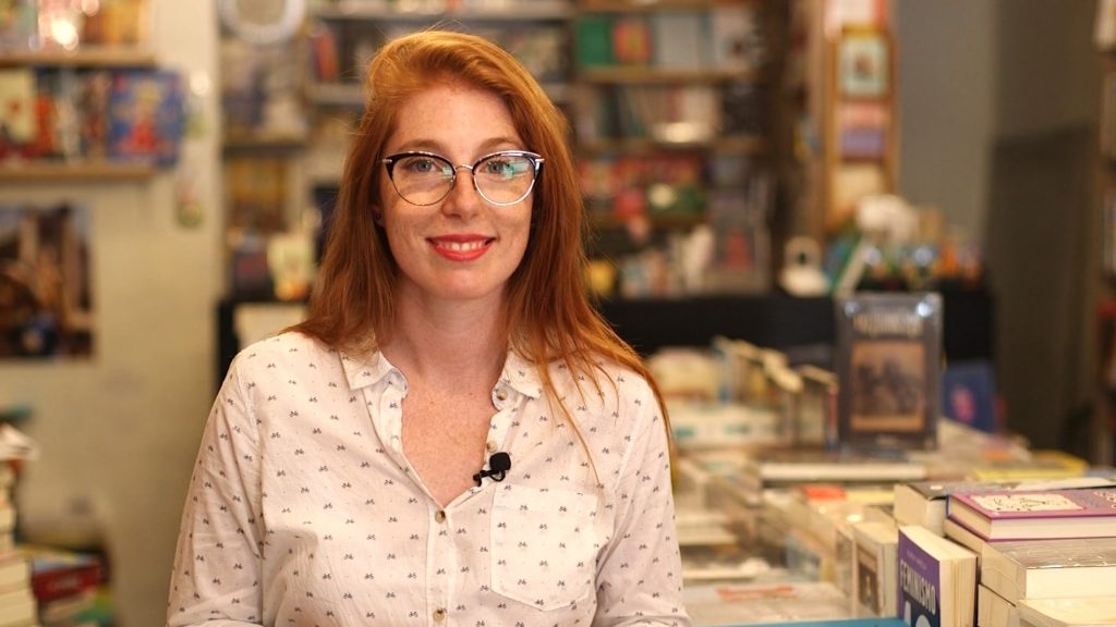 Mujer de cabello rojizo y lentes, sonriendo frente a estantería con libros de fondo en ambiente de biblioteca