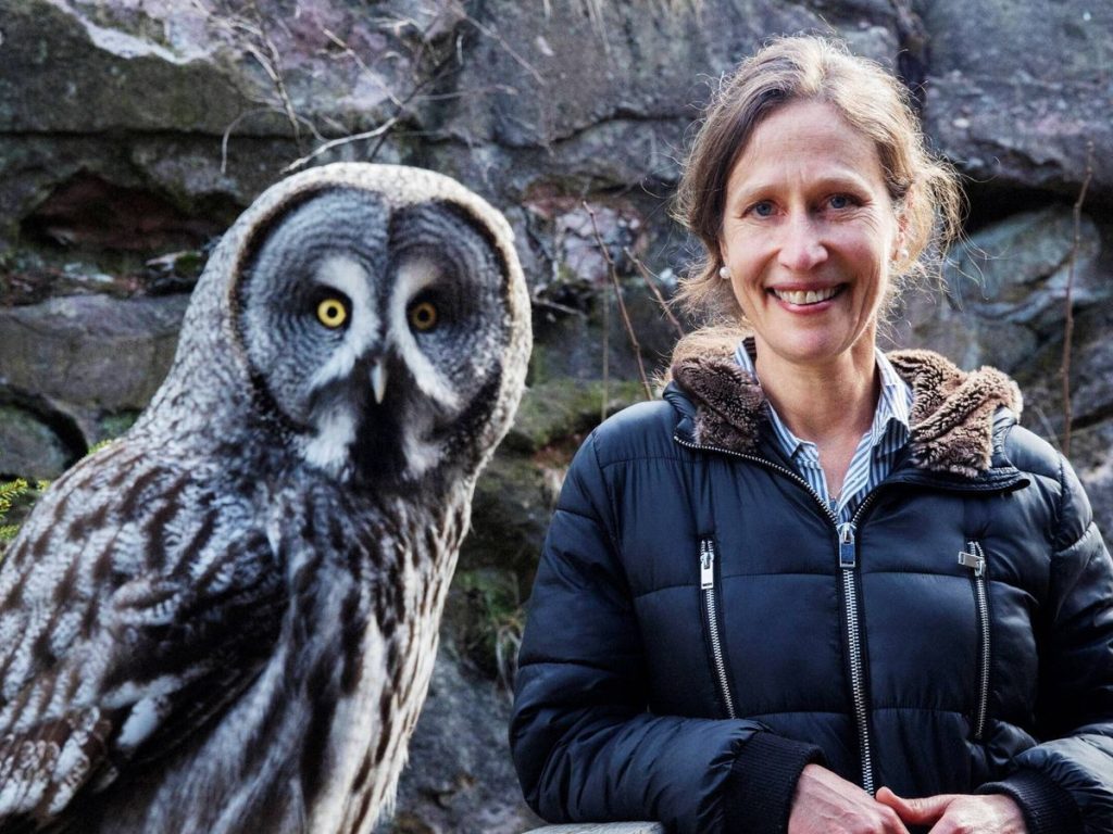 Jennifer Ackerman junto a un búho gris, ambos frente a una pared de piedra natural