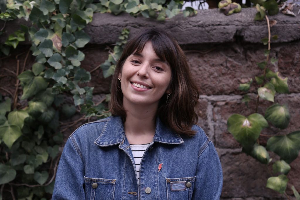 Malú Furche, escritora chilena, sonríe ante una pared de ladrillos con hiedra, vistiendo chaqueta de mezclilla