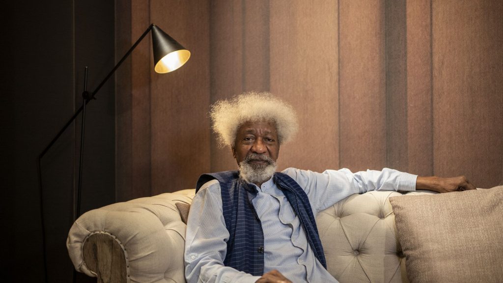 Wole Soyinka con afro blanco, brazos extendidos, sentado en sofá beige bajo luz cálida de lámpara