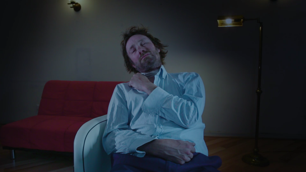 Hombre de mediana edad en camisa azul sentado en sofá rojo, mirando hacia arriba en ambiente oscuro e introspectivo