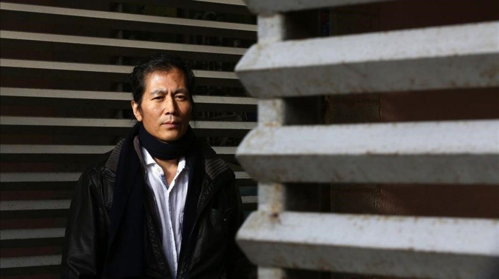 Byung-Chul Han, filósofo surcoreano, en retrato de perfil tres cuartos con blazer negro sobre fondo de líneas grises