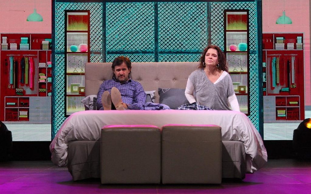 Dos mujeres sentadas en un sofá gris frente a una escenografía colorida con muebles rojos y paneles verdes, en ambiente