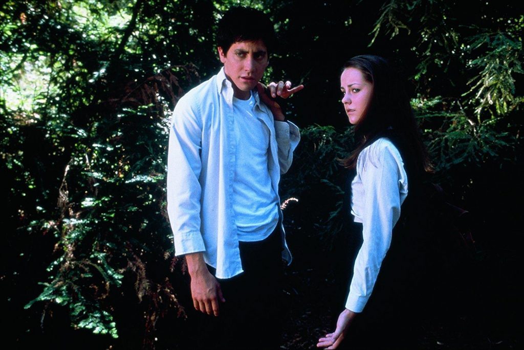 Dos personajes en un bosque nocturno: hombre con camisa azul clara y mujer de cabello oscuro en ropa oscura
