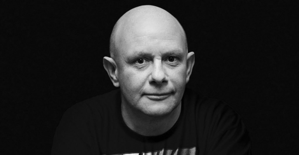 Retrato en blanco y negro de Nick Hornby, escritor británico, contra fondo oscuro, con expresión serena