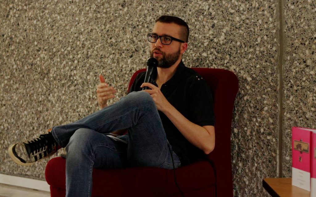 Escritor Luis Panini sentado en sillón rojo, gesturando durante entrevista, con gafas y fondo de papel tapiz