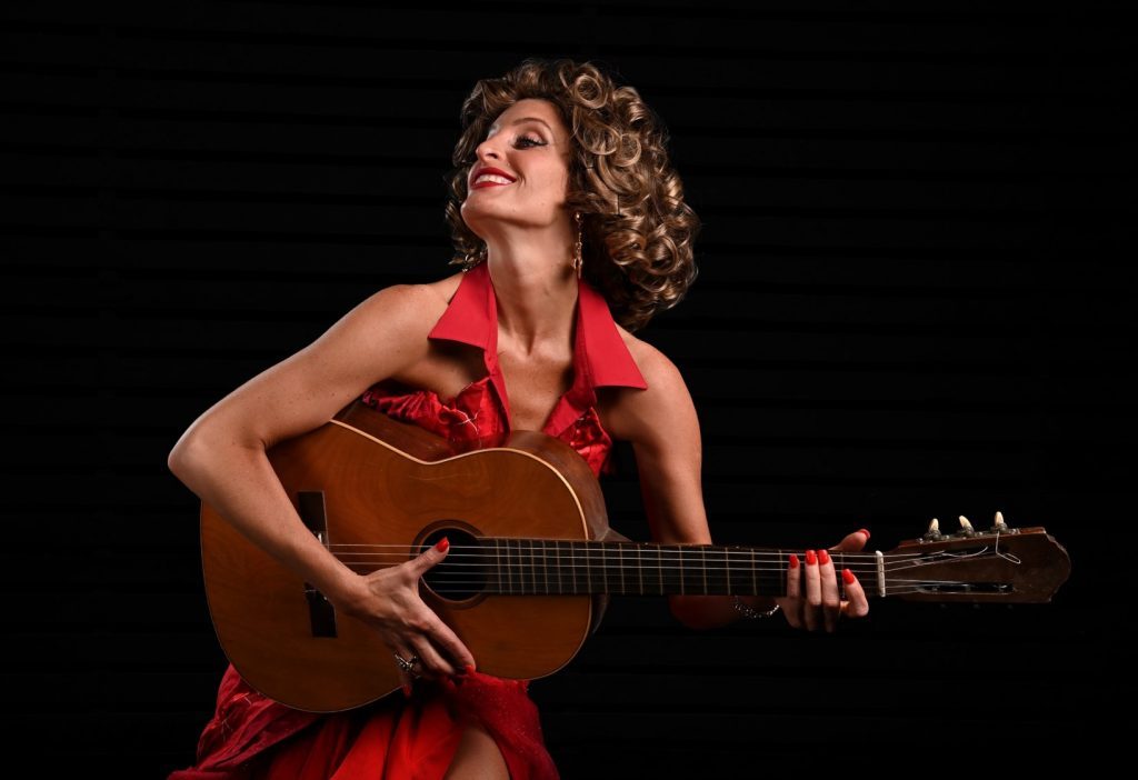 Mujer con cabello rizado en vestido rojo sosteniendo guitarra acústica contra fondo negro