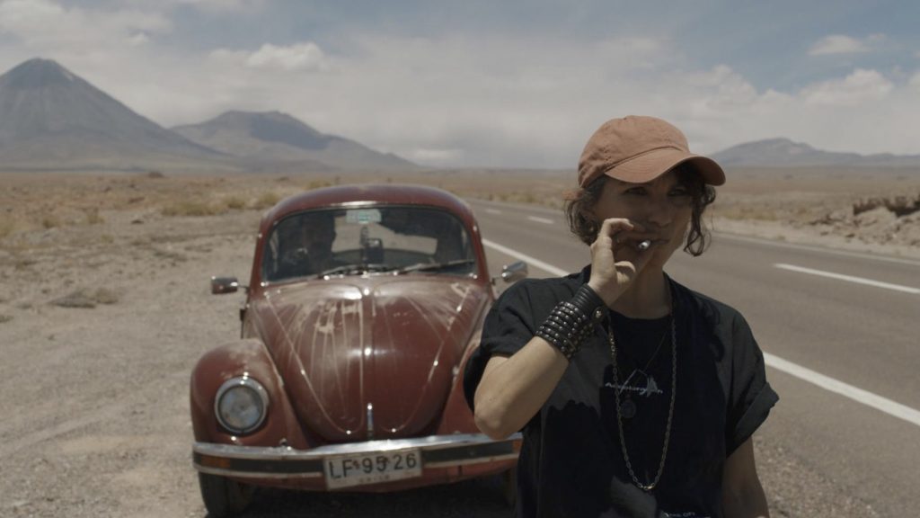 Hombre con gafas de sol junto a un Volkswagen Beetle rojo oxidado en carretera desértica con montañas al fondo