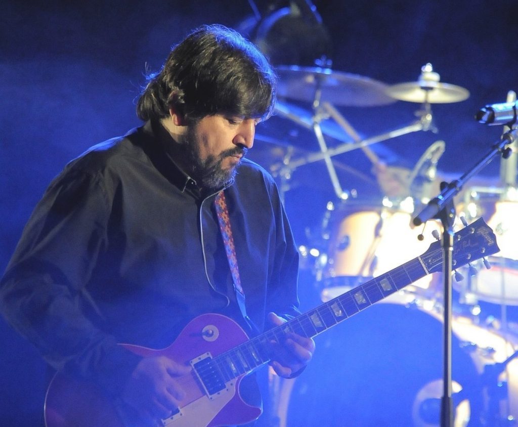 Músico toca guitarra en escenario iluminado con tonos azules, batería de fondo y micrófono en primer plano
