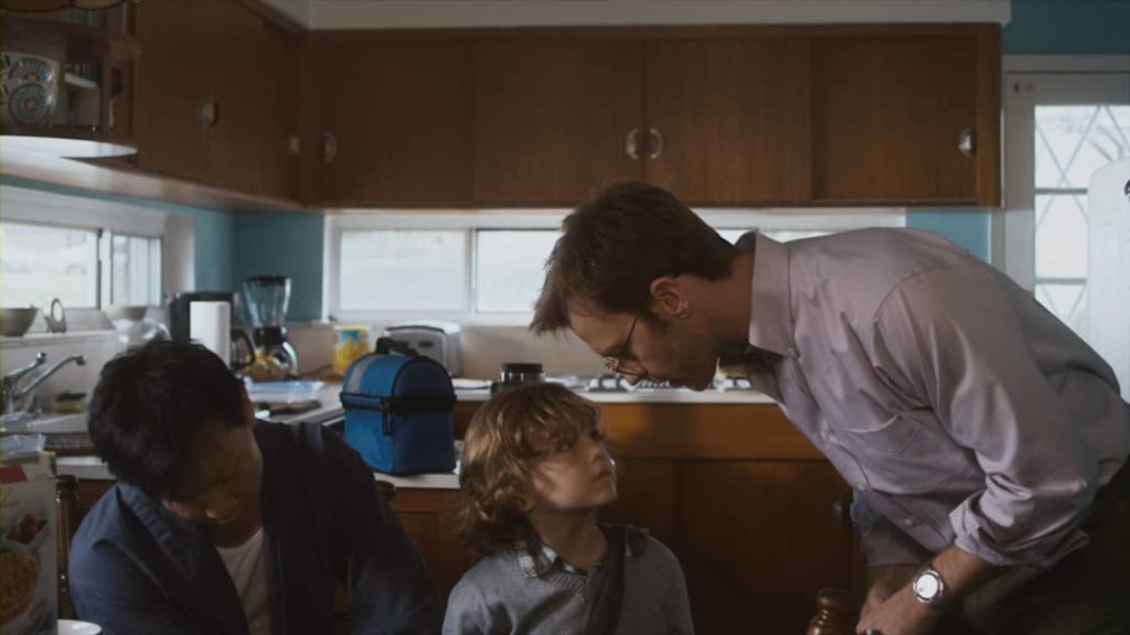 Escena de interior doméstico con tres personas; un adulto se inclina hacia dos niños pequeños en una cocina de madera