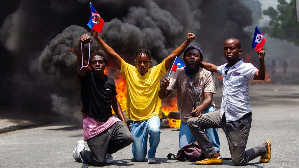 Cuatro personas sostienen bandera haitiana frente a barricada en llamas durante protesta callejera