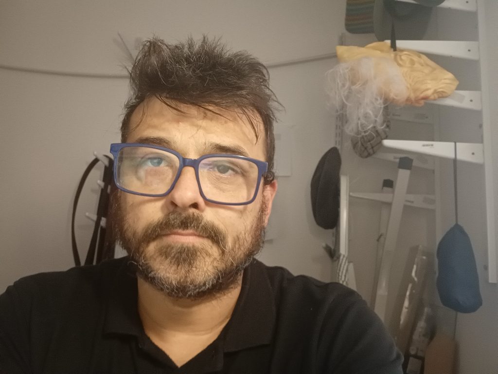 Hombre con gafas y barba oscura, vistiendo polo negro, en interior con equipos de audio de fondo