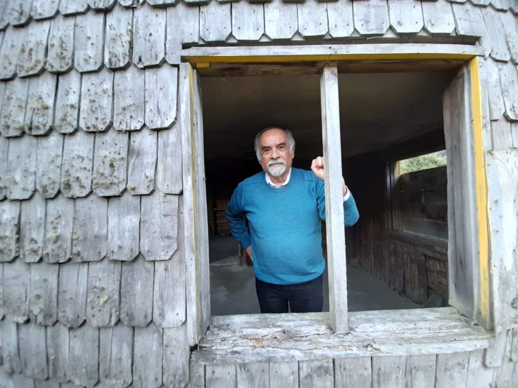 Juan Mihovilovich asomado a ventana de casa rústica con paredes de madera y tejas grises