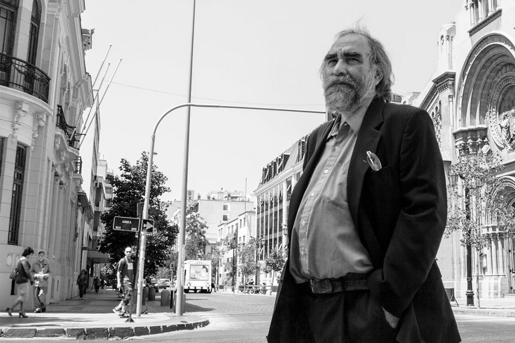 Roberto Merino en calle urbana, traje oscuro y gafas, edificios históricos de fondo, blanco y negro