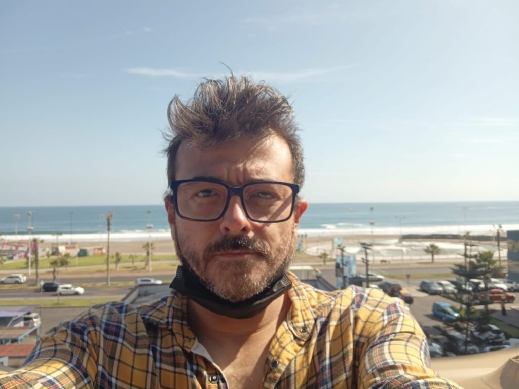 Hombre con gafas y camisa a cuadros amarilla sonriendo ante playa y cielo despejado
