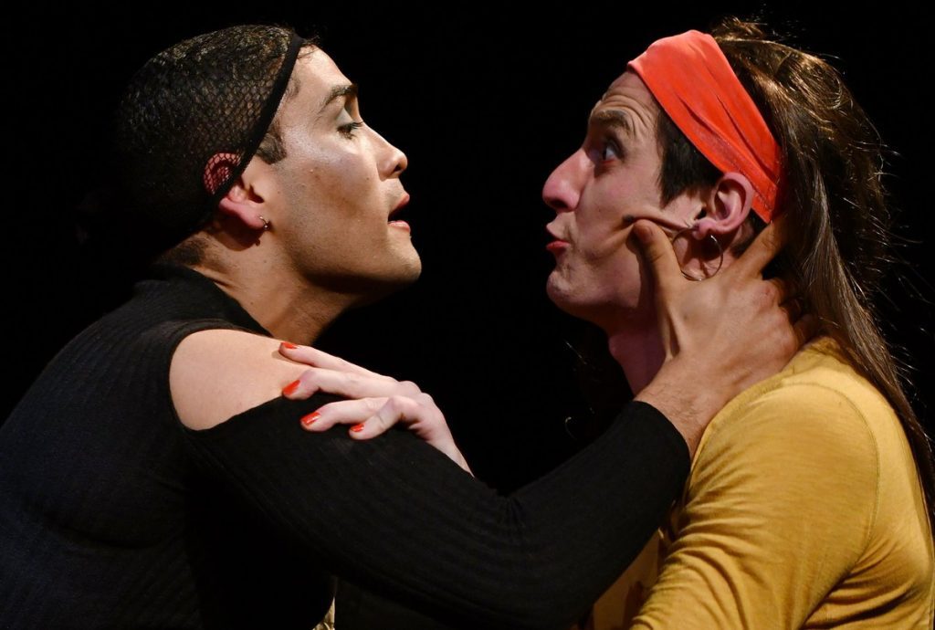Dos hombres frente a frente en escena de teatro, uno de negro y otro de amarillo con pañuelo rojo, en confrontación