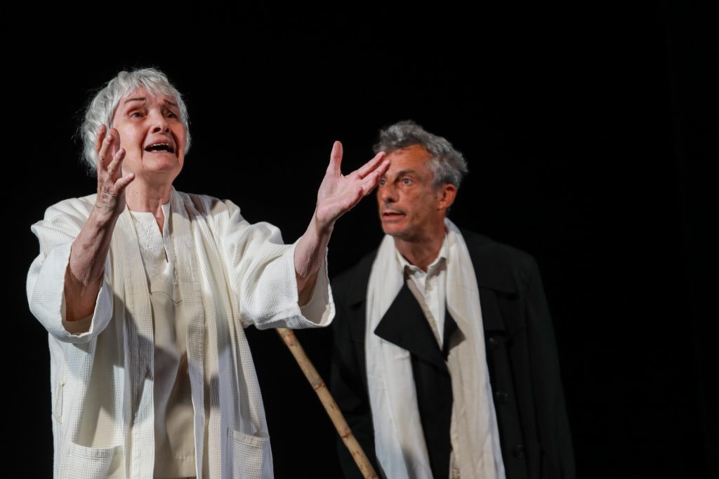 Dos actores en escena representando obra teatral clásica, el de la izquierda con gesto dramático expresivo y capa blanca